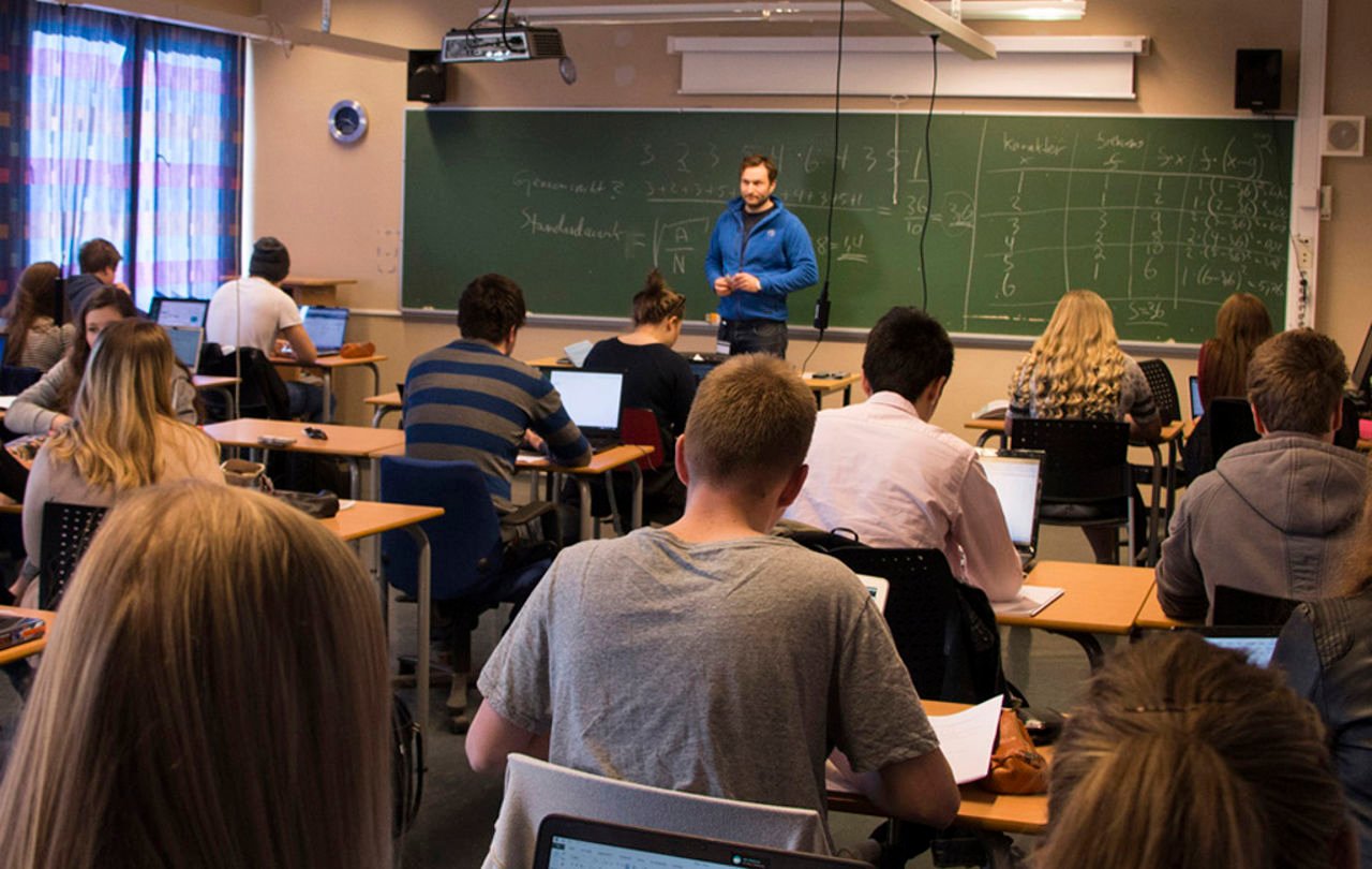 Elevar i eit klasserom, i den vidaregåande skulen. Vi ser ryggane på elevane, og ein blåkledd lærar fremst ved tavla.Foto: Martha Linnea Pukallus.